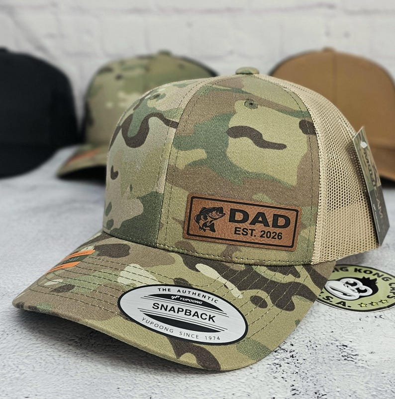 May include: Camouflage print Snapback hat with a brown leather patch that reads "DAD EST. 2026" and a fish graphic. The hat has a tan mesh back and a curved brim. The hat has a white tag that reads "THE AUTHENTIC SNAPBACK YUPOONG SINCE 1974".