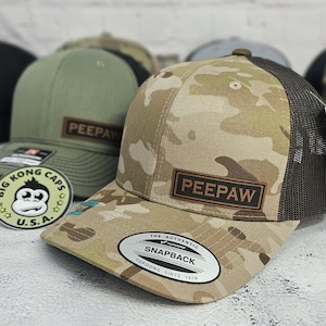 May include: A camouflage print snapback hat with a brown mesh back and a brown leather patch that reads "PEEPAW". The hat has a curved brim and a white tag that reads "SNAPBACK". A green hat with a brown leather patch is visible in the background.