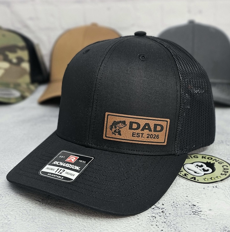 May include: Black trucker hat with a mesh back and a curved brim. A rectangular patch on the front displays the word "DAD" with a fish graphic and "EST. 2026". The hat has an adjustable strap and a Richardson tag.