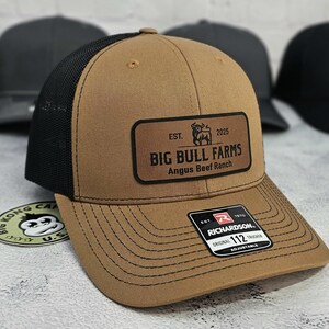 May include: A tan trucker hat with a black mesh back and a curved brim. The hat features a rectangular leather patch with the text "BIG BULL FARMS Angus Beef Ranch" and a bull graphic. The hat is adjustable.
