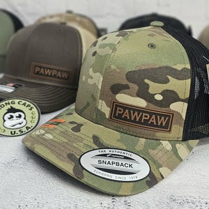 May include: A camouflage pattern baseball cap with a black mesh back and a brown leather patch that reads "PAWPAW". The cap has a curved brim and a snapback closure. The tag reads "THE AUTHENTIC SNAPBACK YUPOONG SINCE 1974".