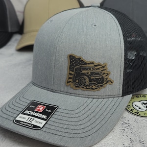 May include: A gray trucker hat with a black mesh back and a curved brim. The hat features a patch with a jeep graphic and American flag design. The Richardson brand label is visible on the front.