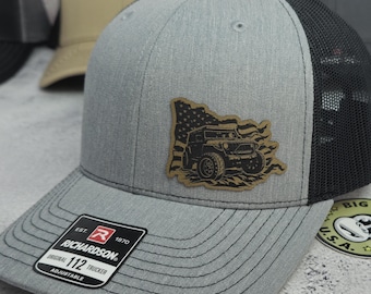 May include: A gray trucker hat with a black mesh back and a curved brim. The hat features a patch with a jeep graphic and American flag design. The Richardson brand label is visible on the front.