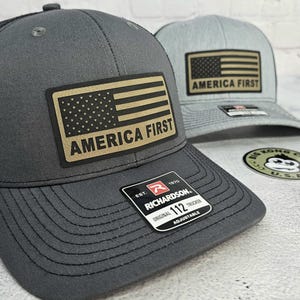 May include: Two trucker hats with the American flag and the text "AMERICA FIRST". The hats are gray and light gray, with a black mesh back. The hats have a patch with the flag design and the text "AMERICA FIRST".