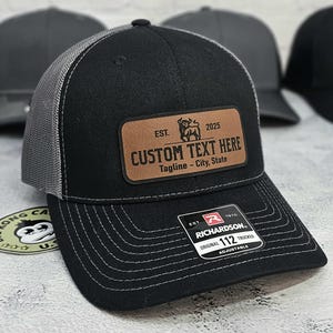 May include: Black trucker hat with a gray mesh back and a brown leather patch. The patch reads "CUSTOM TEXT HERE" with a bull graphic and the text "Tagline - City, State". The hat is adjustable.