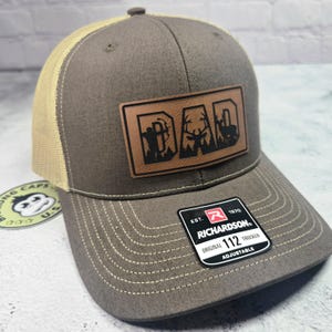 May include: A brown and tan trucker hat with a leather patch that says "DAD" with a hunting theme. The patch features a hunter with a bow, a deer, and a silhouette of a deer. The hat has a curved brim and a mesh back.