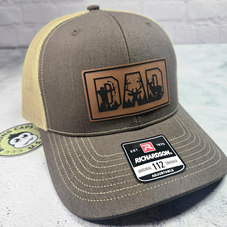 May include: A brown and tan trucker hat with a leather patch that says "DAD" with a hunting theme. The patch features a hunter with a bow, a deer, and a silhouette of a deer. The hat has a curved brim and a mesh back.
