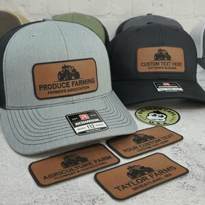May include: Gray and black trucker hat with a brown leather patch featuring a tractor and the text "PRODUCE FARMING FARMER'S ASSOCIATION." A black hat with a similar patch reads "CUSTOM TEXT HERE CITY STATE SLOGANS."