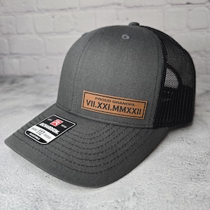 May include: A gray and black trucker hat with a brown leather patch that says "Proud Grandpa VII.XXI.MMXXII".