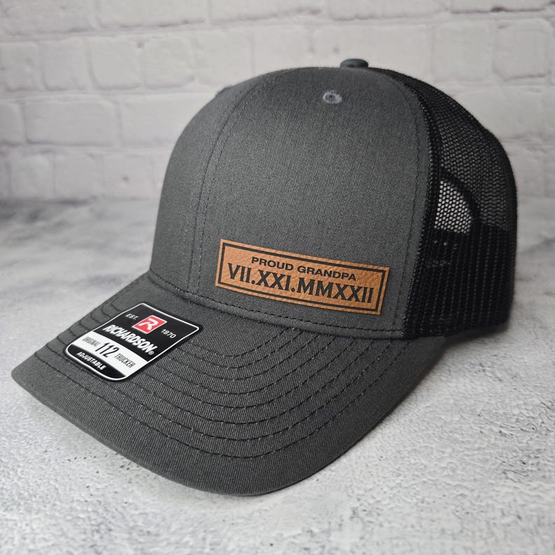 May include: A gray and black trucker hat with a brown leather patch that says "Proud Grandpa VII.XXI.MMXXII".