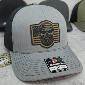 May include: Gray trucker hat with a black mesh back and a patch featuring a skull with an American flag design. The hat has a curved brim and the brand name "Richardson" is visible on a tag.