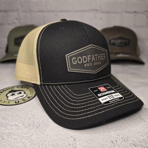 Puede incluir: Gorra de camionero negra y beige con un parche de cuero marr&oacute;n que dice "Godfather Est. 2025". La gorra es una Richardson 112 ajustable.