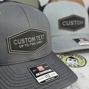 May include: Close-up of two gray trucker hats with a custom text patch. The patch reads "CUSTOM TEXT UP TO TWO LINES." One hat has a black patch that says "CUSTOM." The hats have a Richardson 112 Trucker tag.