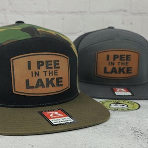 May include: Two flat bill snapback hats. One hat has a camouflage pattern on the top and a brown brim. The other hat is gray. Both hats have a brown leather patch with the text "I PEE IN THE LAKE".
