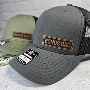 May include: Two trucker hats, one olive green and one gray, with a mesh back. Each hat has a rectangular patch that reads "BONUS DAD" in brown lettering. The hats are adjustable and have a Richardson 112 Trucker tag.