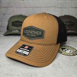 Puede incluir: Una gorra de camionero marr&oacute;n y negra con una malla negra en la parte posterior. La parte delantera de la gorra tiene un parche verde oscuro con el texto "GODFATHER EST. 2025". La gorra est&aacute; hecha por Richardson 112.