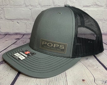 Puede incluir: Una gorra de camionero gris y negra con un parche marrón que dice "POPS ESTABLISHED 2024".
