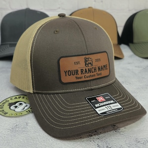 May include: A brown trucker hat with a tan mesh back. The hat features a leather patch with the text "YOUR RANCH NAME" and "Your Custom Text". The patch also includes a bull graphic and the year "2025". The hat is adjustable.