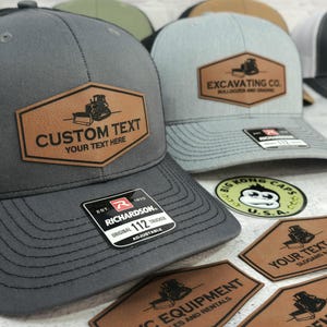 May include: Gray and light blue trucker hats with brown leather patches. One patch reads "CUSTOM TEXT YOUR TEXT HERE" and the other reads "EXCAVATING CO. BULLDOZER AND GRADING". The hats are adjustable and have a Richardson 112 tag.