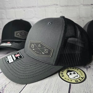 May include: A gray and black trucker hat with a black mesh back. The hat has a gray patch on the front with the letters "B & S" and the date "08.24.25" in white. The hat is sitting on a wooden surface with a sticker that says "Big Kong Caps U.S.A."