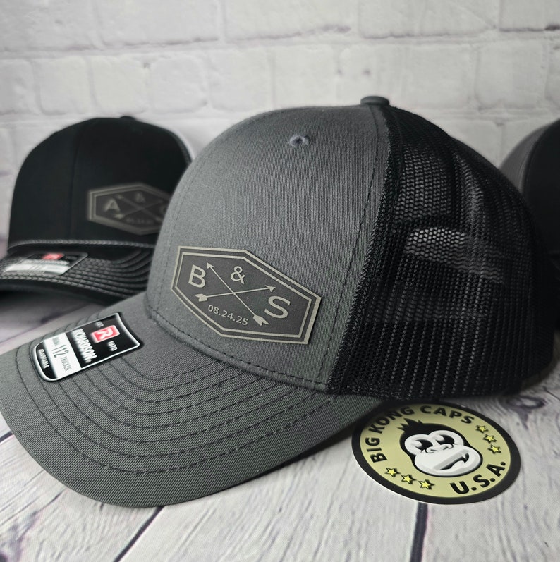 May include: A gray and black trucker hat with a black mesh back. The hat has a gray patch on the front with the letters "B & S" and the date "08.24.25" in white. The hat is sitting on a wooden surface with a sticker that says "Big Kong Caps U.S.A."