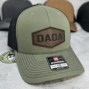 May include: Olive green trucker hat with a black mesh back and a brown leather patch that reads "DADA". The hat is a Richardson 112 Original Trucker, adjustable.