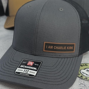 May include: A gray Richardson 112 trucker hat with a black mesh back. The hat features a brown leather patch with the text "I AM CHARLIE KIRK". The hat is adjustable.