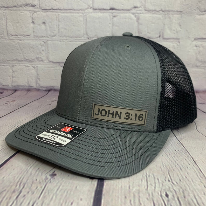 May include: A gray and black trucker hat with a brown leather patch that says "JOHN 3:16".