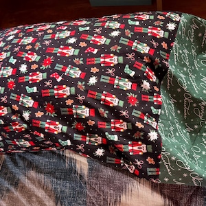 May include: A rectangular pillow with a black background, patterned with red, white, and green nutcrackers, snowflakes, and bows. The pillow has a green border with the words "Merry Christmas" in white script.