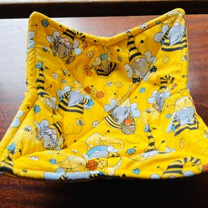 May include: A yellow quilted hot pad with a bee and gnome pattern. The hot pad is folded in half and has a square shape.
