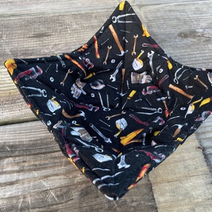 May include: A black quilted hot pad with a pattern of various tools, including hammers, wrenches, screwdrivers, and nails. The hot pad is square shaped and has a quilted texture.