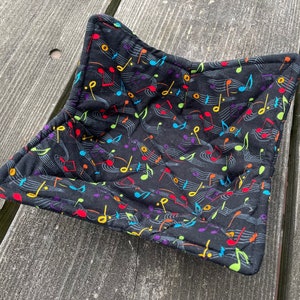 May include: Black fabric bowl with a colorful musical note pattern. The bowl is made of quilted fabric and has a square shape.