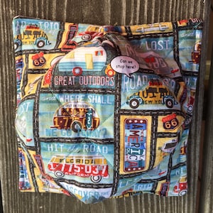 May include: A quilted fabric pouch with a vintage travel theme. The pouch features a variety of images, including a camper van, a car, and a road sign that says "Can we stop here?" The pouch is decorated with the words "Great Outdoors", "Road Trip", and "Hit the Road".