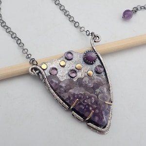 May include: A triangular pendant necklace featuring a textured silver frame with small gold accents and embedded purple amethyst stones. The pendant hangs from a dark silver chain, which includes a single purple bead near the clasp closure.