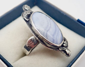 May include: A silver ring with a blue agate stone set in a decorative oval bezel. The ring is in a white box with a blue lining.