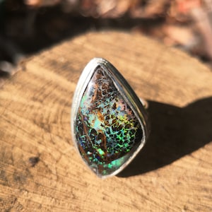 May include: A silver ring with a large, iridescent opal gemstone. The opal has a dark brown base with green, blue, and purple flashes of color.