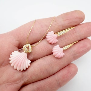 May include: A gold necklace and earrings set featuring pink seashell pendants with clear crystal accents. The necklace has a heart-shaped pendant.