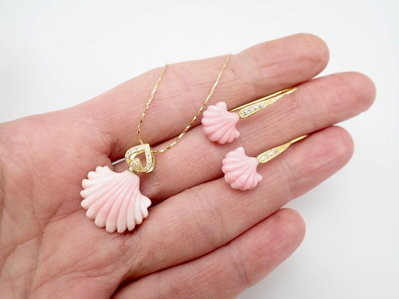 May include: A gold necklace and earrings set featuring pink seashell pendants with clear crystal accents. The necklace has a heart-shaped pendant.