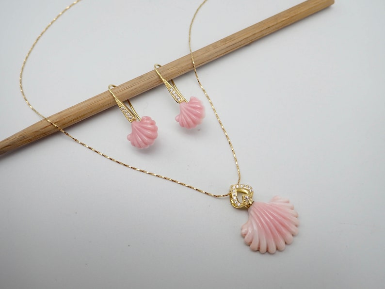 May include: A gold necklace and earrings set featuring pink shell pendants. The necklace has a delicate chain and the earrings have a drop design.