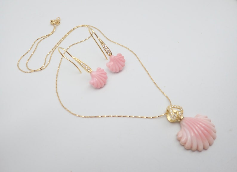 May include: A delicate gold chain necklace with a pink seashell pendant and matching earrings. The pendant and earrings are adorned with small clear crystals.