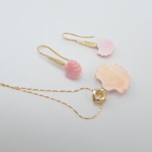 May include: A gold necklace with a pink seashell pendant and matching earrings. The pendant is shaped like a seashell and has a heart-shaped cutout in the center. The earrings are also shaped like seashells and have a small gold bead at the top.