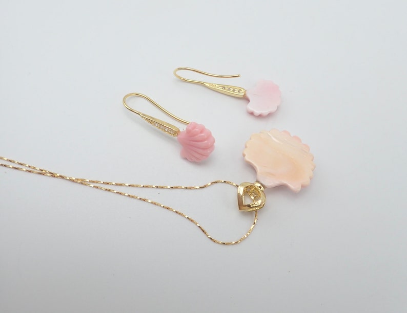 May include: A gold necklace with a pink seashell pendant and matching earrings. The pendant is shaped like a seashell and has a heart-shaped cutout in the center. The earrings are also shaped like seashells and have a small gold bead at the top.