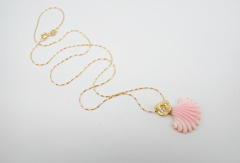 May include: A delicate gold chain necklace with a pink coral shell pendant. The pendant features a small gold charm with clear stones.
