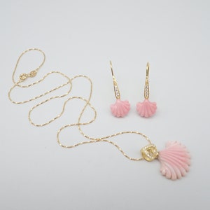 May include: A delicate gold chain necklace with a pink seashell pendant and matching earrings. The seashells have a detailed design and are accented with small clear stones.