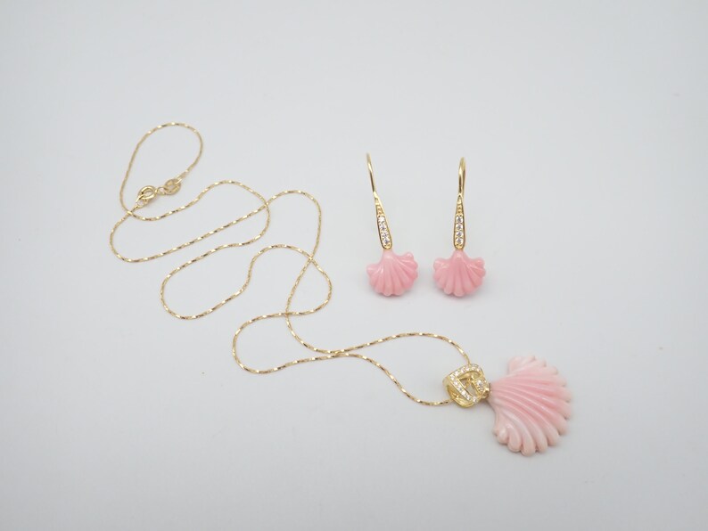 May include: A delicate gold chain necklace with a pink seashell pendant and matching earrings. The seashells have a detailed design and are accented with small clear stones.