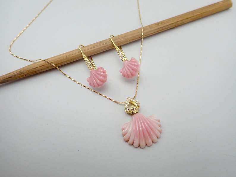 May include: A delicate gold chain necklace with a pink shell pendant accented with small clear stones. Matching pink shell earrings with gold hooks.