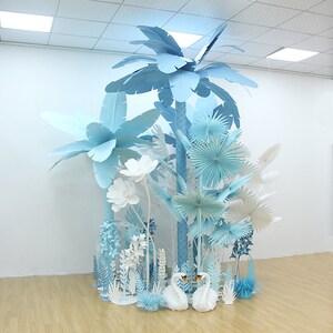 K&ouml;nnte beinhalten: Eine Papierkunstinstallation mit einer blauen und wei&szlig;en tropischen Szene mit Palmen, Blumen und Schw&auml;nen. Die Installation ist aus Papier gefertigt und in einem wei&szlig;en Raum ausgestellt.
