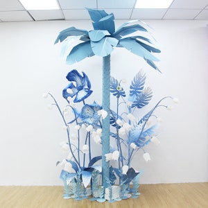 K&ouml;nnte beinhalten: Ein blauer Papier-Palmenbaum mit blauen Papierblumen und Bl&auml;ttern in einem wei&szlig;en Raum. Die Blumen sind in verschiedenen Blaut&ouml;nen und Wei&szlig;.