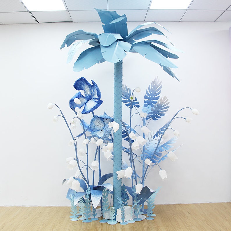 K&ouml;nnte beinhalten: Ein blauer Papier-Palmenbaum mit blauen Papierblumen und Bl&auml;ttern in einem wei&szlig;en Raum. Die Blumen sind in verschiedenen Blaut&ouml;nen und Wei&szlig;.
