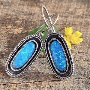 Peut inclure: Une paire de boucles d'oreilles en argent avec des pierres d'opale bleues serties dans un cadre ovale noir. Les boucles d'oreilles ont un design simple et élégant.